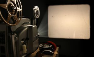 A vintage 8mm film projector projects a blank image with film dust and scratches onto a wall beside a stack of film reels