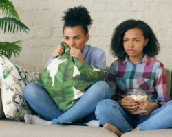Zwei Jugendliche sitzen mit Popcorn und Kissen gemeinsam auf dem Sofa